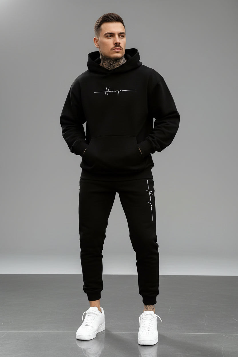 Man wearing a black hoodie and pants with a logo on a street.