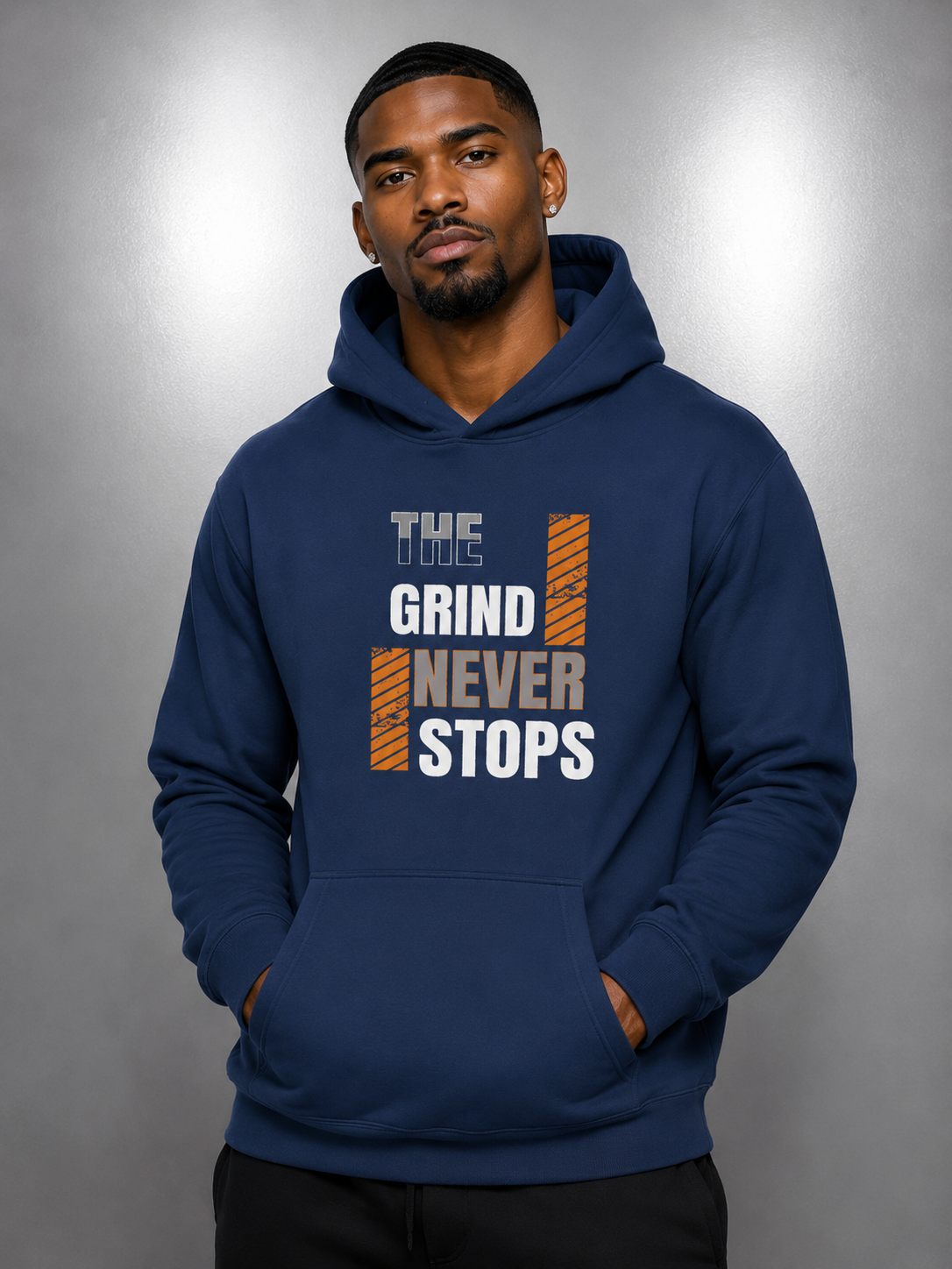 A man wearing a navy blue pullover hoodie with a graphic print on the chest that reads 'THE TOYS GRIND NEVER STOPS' in white and orange, posed against a gray studio background.