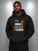 A man wearing a black pullover hoodie with a graphic print that reads 'THE GRIND NEVER STOPS' in white and orange text, posed against a gray studio background.