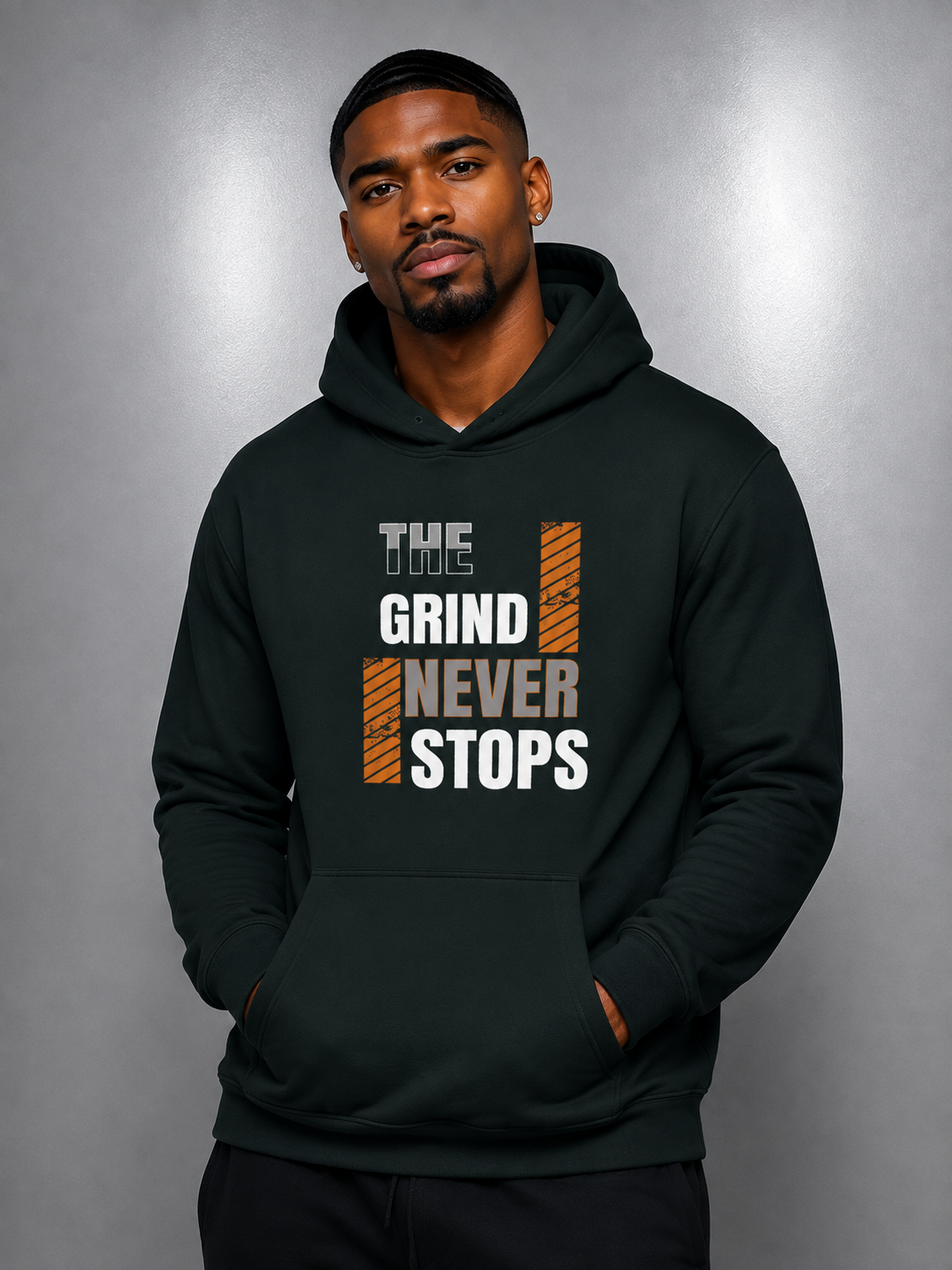 A man wearing a dark forest green hoodie with a graphic print that reads 'THE GRIND NEVER STOPS' in white and orange, posed against a gray studio background.