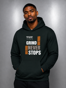 A man wearing a dark forest green hoodie with a graphic print that reads 'THE GRIND NEVER STOPS' in white and orange, posed against a gray studio background.